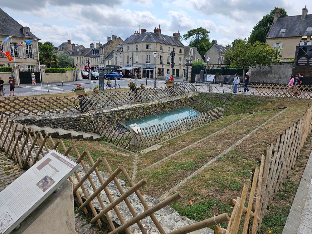 A sunken grassy area in the middle of a city, surrounded by a fence. At the bottom is a rectangular area covered by glass