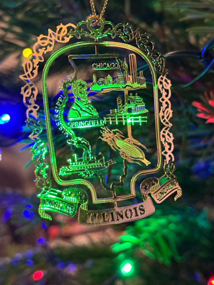 A metal Christmas ornament depicting scenes from Illinois, including a river boat, a barn, the likeness of Abraham Lincoln, the skyline of Chicago and, yes, an ear of goddamn corn.