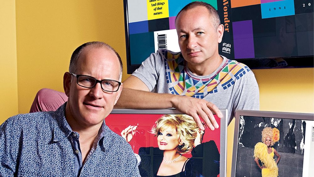Portrait of Fenton Bailey, pictured right, and Randy Barbato, pictured left, sitting at a desk with two computer screens against a yellow wall.