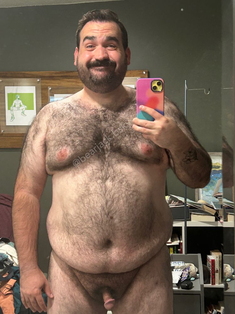 A hairy naked man with dark hair and a mustache and scruff stands in front of a mirror holding an iPhone