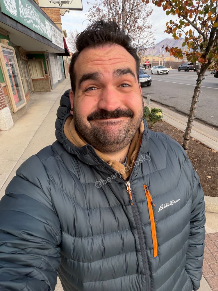 A man with dark hair and a mustache walking outside on a city sidewalk. He is wearing a dark blue puffer jacket 