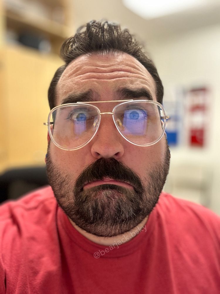 A man with dark hair, a mustache, and short beard wearing large gold-framed glasses and a red shirt makes an exaggeratedly large frown and pouty eyes 