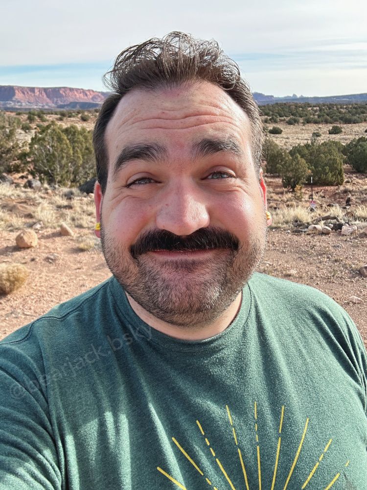 A man with dark hair and a mustache wearing a green tshirt stands in the desert with red rocks in the background 