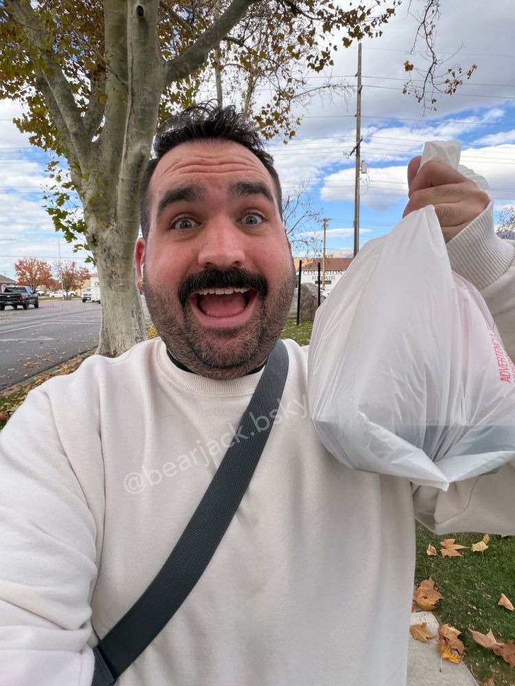A man in a cream sweatshirt holds up a bag of food and smiles with an open mouth