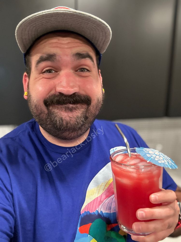 A man in a blue shirt and baseball cap with a beard and mustache. He’s holding a glass with a red drink, a metal straw, and a blue umbrella 