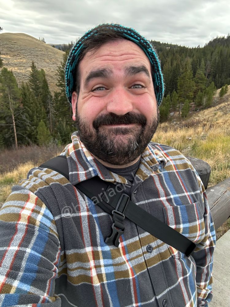 A man with dark hair and a beard standing outside with evergreen trees in the background. He is wearing an electric blue and charcoal colored beanie, and a charcoal gray plaid flannel shirt. He is smiling 