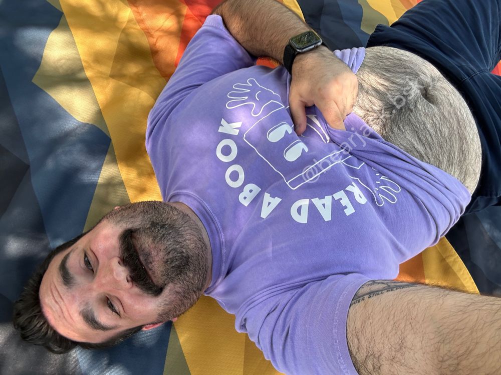A man with dark hair, a mustache, and scruff lying down on a blanket lifting up his light purple shirt to show his hairy stomach 