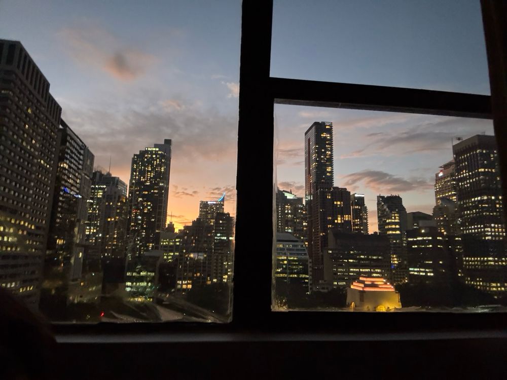 View of Sydney city buildings. A pale orange sunset glows behind the cityscape, and hundreds of windows glow with lights. In the foreground on the right, the illuminated ANZAC memorial is almost the same colour as the setting sun. 