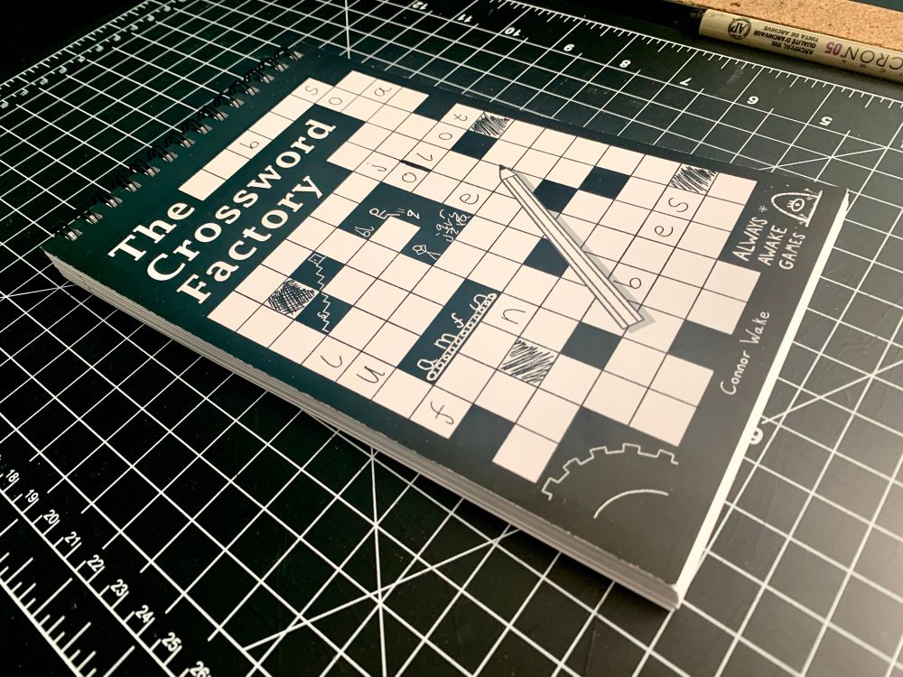 A spiral bound book with the title The Crossword Factory and a sporadically filled in crossword on the cover. There are little stick figures working in a letter factory in the blocks of the crossword. 