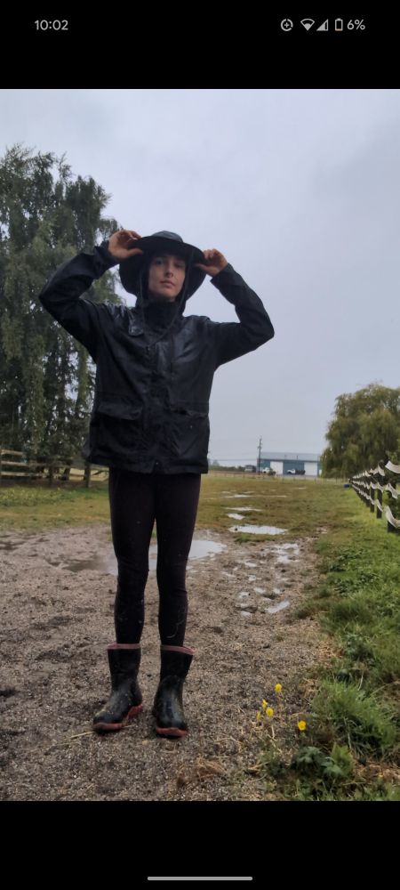 Josie decked out in black rain gear standing in a field with many puddles about. 
