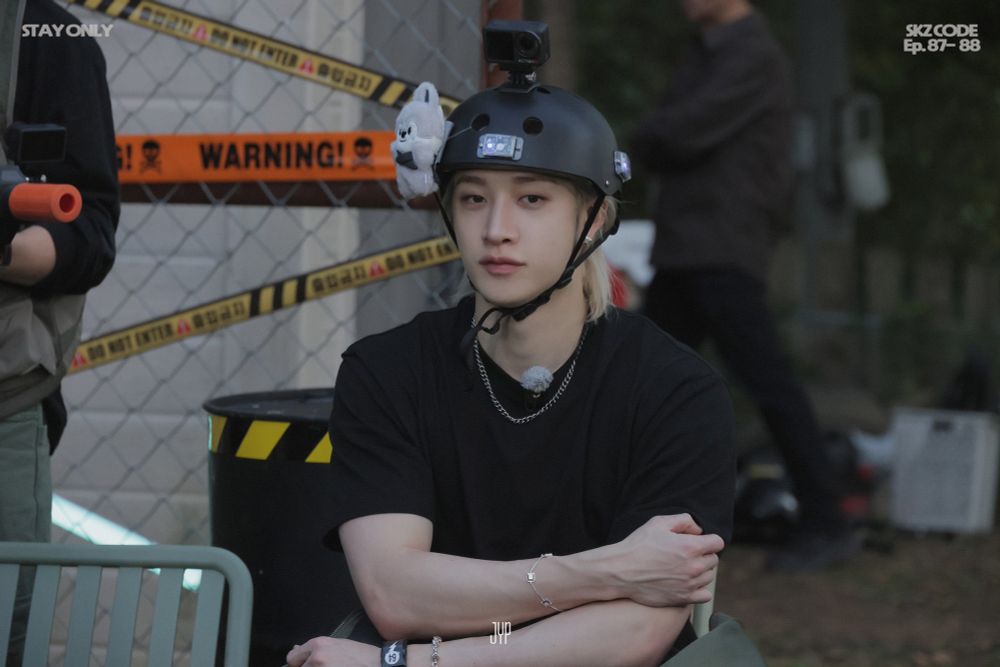 Chan sitting in a chair. He’s wearing a black shirt, and a helmet with a Wolf chan in it. It’s a behind the scenes photo of ep. 87 and 88 of SKZ Code.