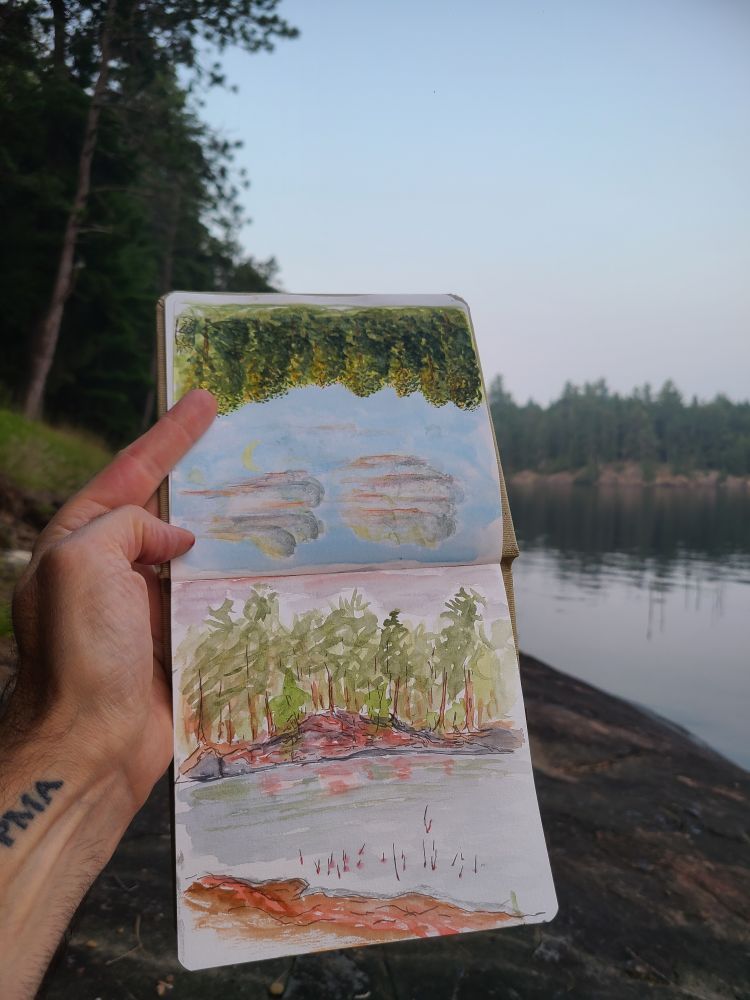 A sketchbook is held up in front of the scene depicted. Candian sheild cliff on Jean Island catches the red light of a sun setting through wildfire smoke. Som reeds break the surface in the foreground. May all beings find a peace that is independent of circumstances ✌️ 