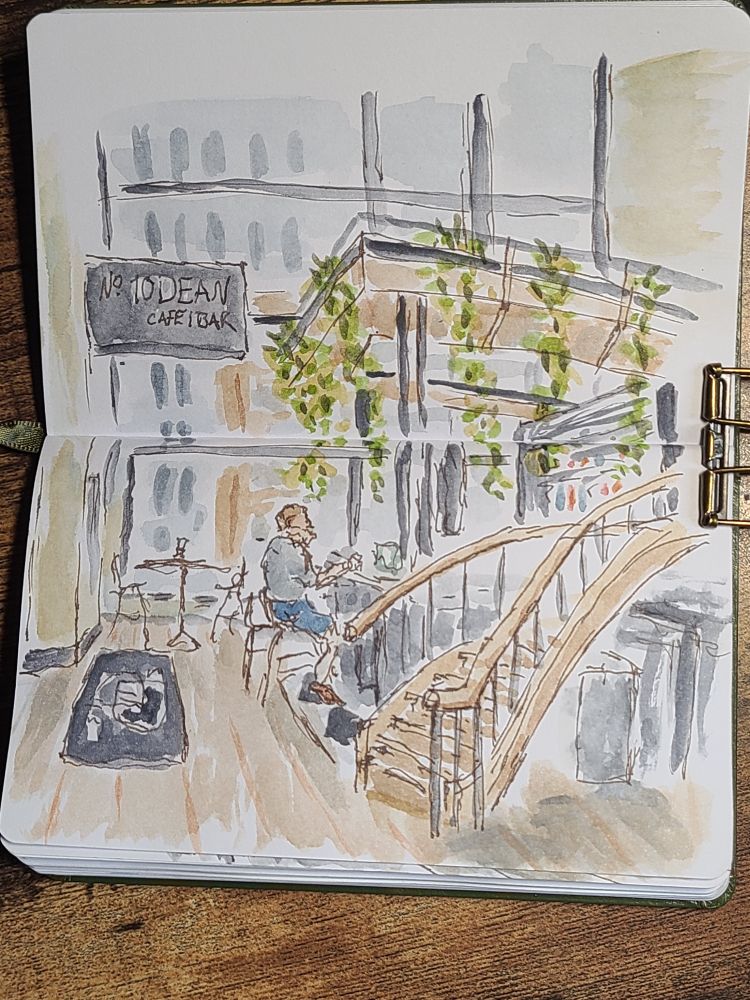 An open sketchbook shows a scene of a light, airy cafe into which a curving staircase descends. Someone sits at the bar, and some buildings are visible through the large windows. Did you read this? I spend a fair amount of time and effort on alt-text and want to know if it's reaching anyone. Let me know in the comments? Have a lovely day!