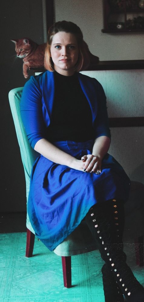 eve sitting down in a chair wearing a blue skirt and a blue crop top blazer with high heels on and her cat behind her