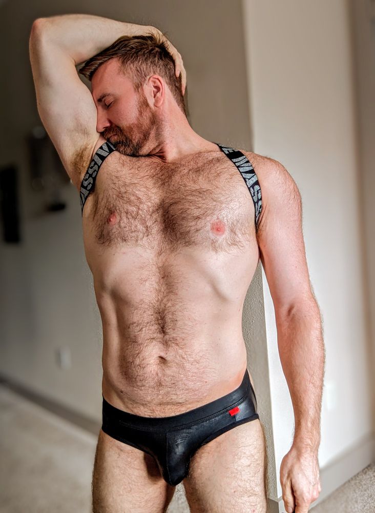 Hairy bearded muscle guy sniffing his armpit while wearing a black leather jockstrap and black chest harness