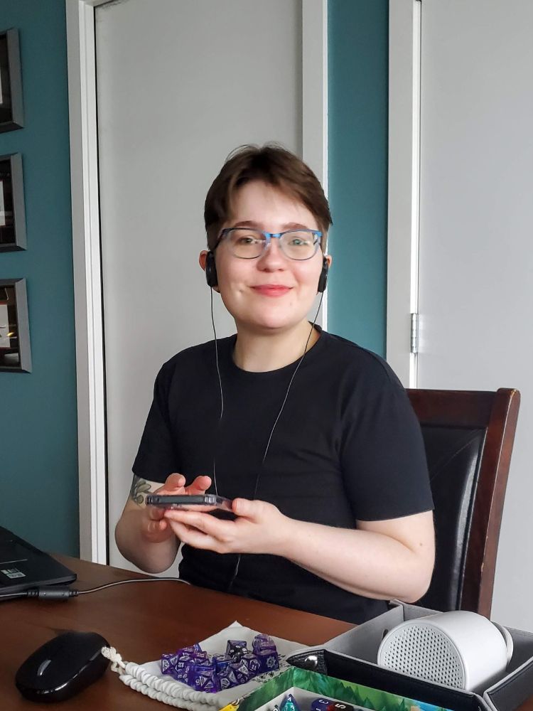 A photo of me, a white métis person sitting at a table with TTRPG paraphernalia in front of me: polyhedral dice, a bluetooth speaker, a laptop. I have my phone in hand and wired headphones on my ears. I have short hair and am wearing eyeglasses and a black t-shirt, and I'm smiling at the camera
