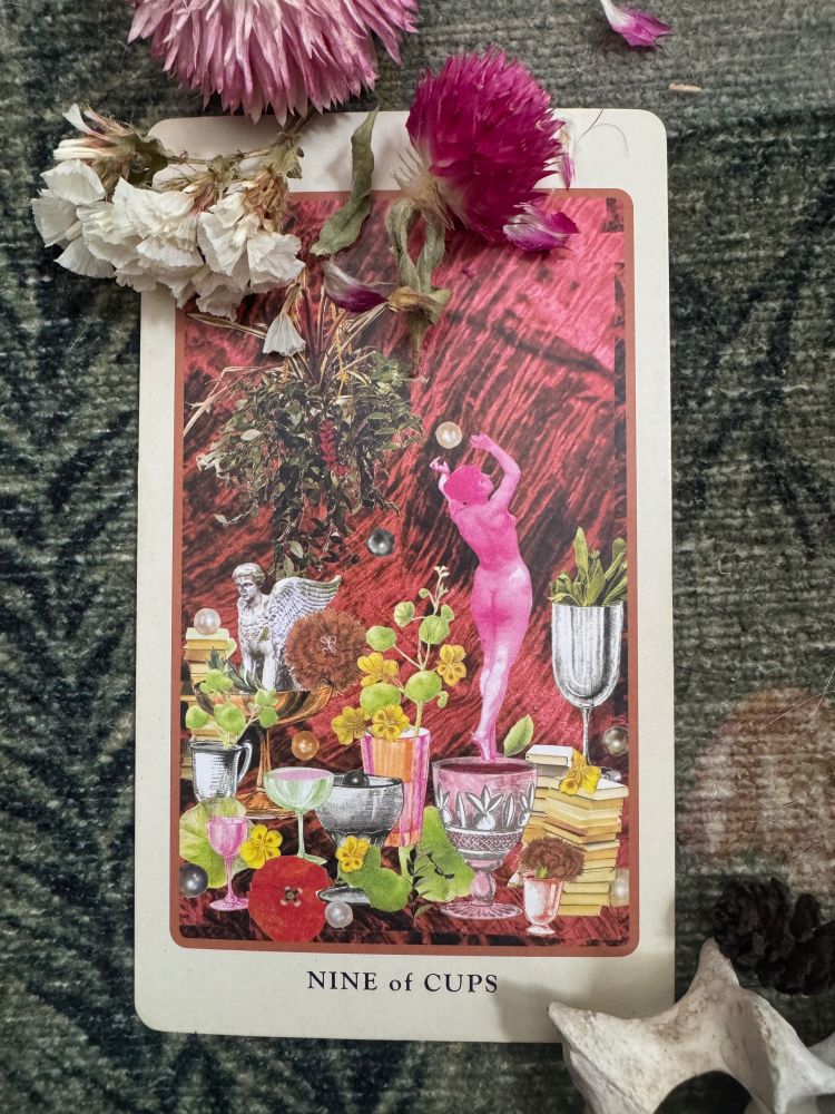 A single tarot card, the nine of cups, with flowers. The card is collaged with colorful images including a hot pink naked lady. on her tiptoes with her back to the viewer and her arms raised. She is poised on the rim of a cup, surrounded by flowers, books, art, and treats. Cups are water in the tarot, the realm of imagination, emotion, and the subconscious. This card can be associated with indulgence. I also associate it with confidence and experience in the realm of cups. The gift of knowing what pleasures fill you particular cups, and perhaps even how to magnetize more of them to you. On this day, a beautiful reminder to tend to your feelings and dreams. 