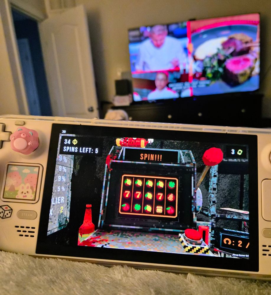A photo of my Steam deck with clover pit on the screen. Hell's Kitchen is on the tv in the background.
