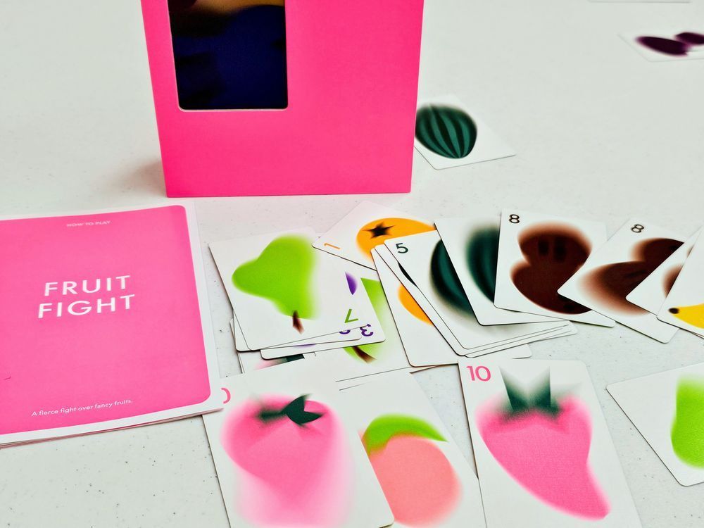 A game from the CMYK Magenta line. The bright pink box and rule book are on the table. Some of the cards are spread out on the table. They depict an artistic look of fruit.