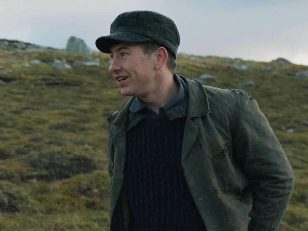 Still photo of Barry Keoghan in profile, as the character Dominic in the movie The Banshees of Inisherin (2022). Behind him rises a rugged mossy landscape and an overcast sky.