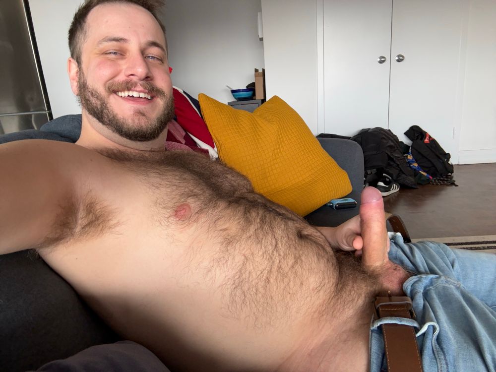 Side-view photo of me sitting shirtless on a couch with my hard cock poking up from my jeans. My hairy pit, chest, belly, and bush are lit up from the sunlight behind the camera.