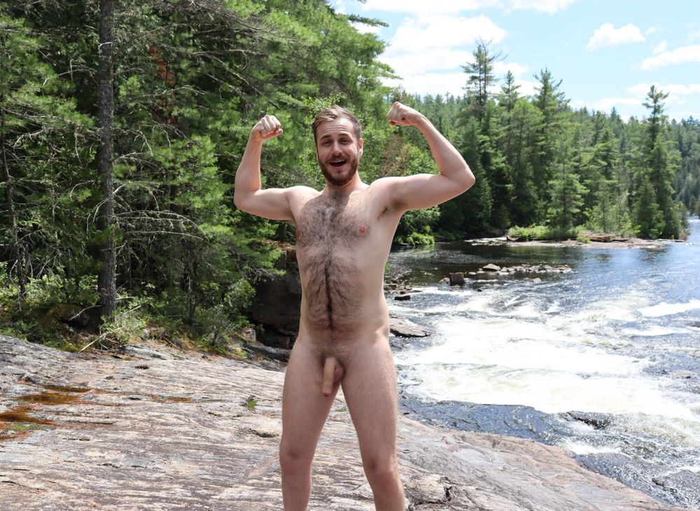 Photo of me naked, flexing, standing on a rock in front of a river and forest in the mountains. The sun is shining behind me, casting a light shadow over my face and body. My soft, uncut cock is hanging, but it’s a bit smaller than normal from the cold air. 