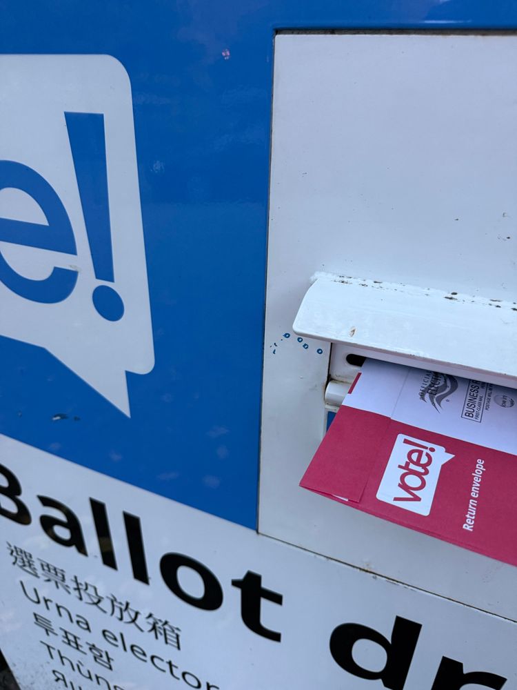 A red and white ballot envelope is being inserted into a white and blue ballot box.