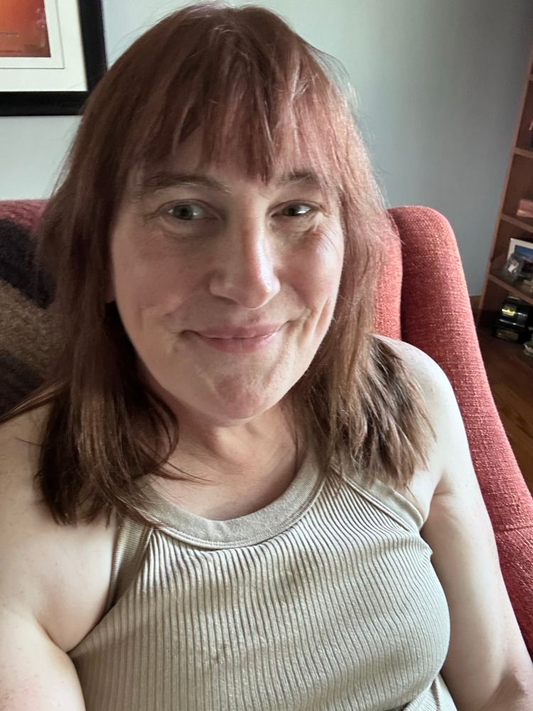 A beautiful white woman sits in a sunny living room on a red orange couch. Her long brown hair falls to the shoulders of her halter style beige shirt. The rim could be lit by her smile as she almost flirts with the camera.