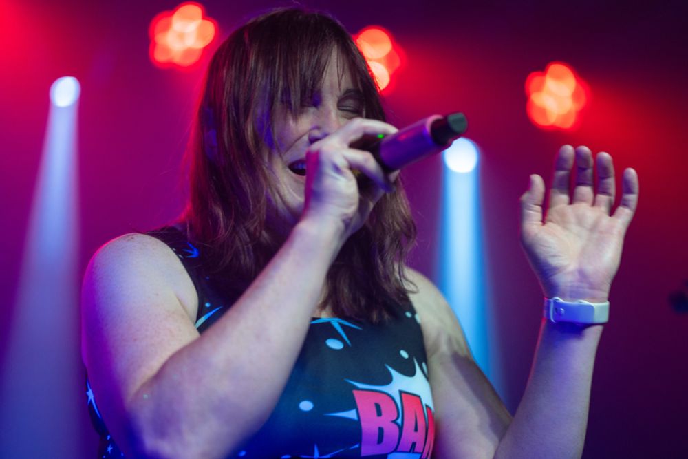 A pretty white woman in her mid 40s stands on a stage performing, backlight with red lights and blue lights, illuminating in the fog. Her long brown hair comes to the shoulders of her pop art space themed dress. She’s in the middle of singing and has a pink wireless microphone up to her mouth with the other hand raised.