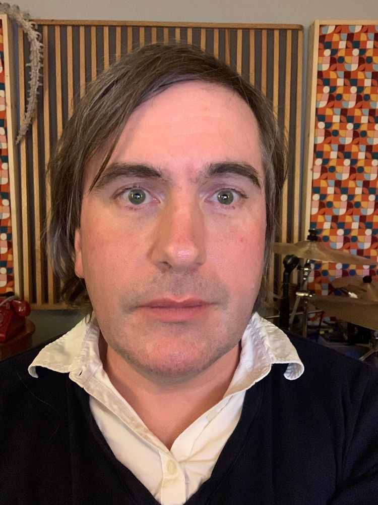 Pre-transition photo of Emily, here appearing as a white man with shortish brown gray hair, looking into the camera with dead eyes. They wear a T-shirt of dark blue with a button up white colored shirt underneath behind them is ornate acoustic treatment on the wall made of a combination of vertical wood slots and colorful acoustic panels.