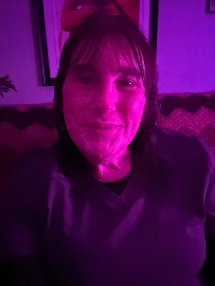 A white woman with long dark hair sits in a dim living room lit up by the glow of a fluorescent Halloween cat unseen in this image. She’s wearing gray scrubs with a black undershirt and looks confidently into the camera.