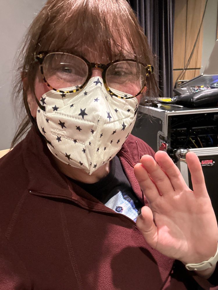 A white woman sits in the back of the theater while all of the sound equipment. She’s turned in an angle waving and we can’t see her whole face because she’s wearing a white face mask with black stars on it and glasses and her long brown hair hold back in a ponytail. She’s wearing a red zip fleece. That’s unzipped enough to see What looks like the eye of a cat peeking out from a black T-shirt.