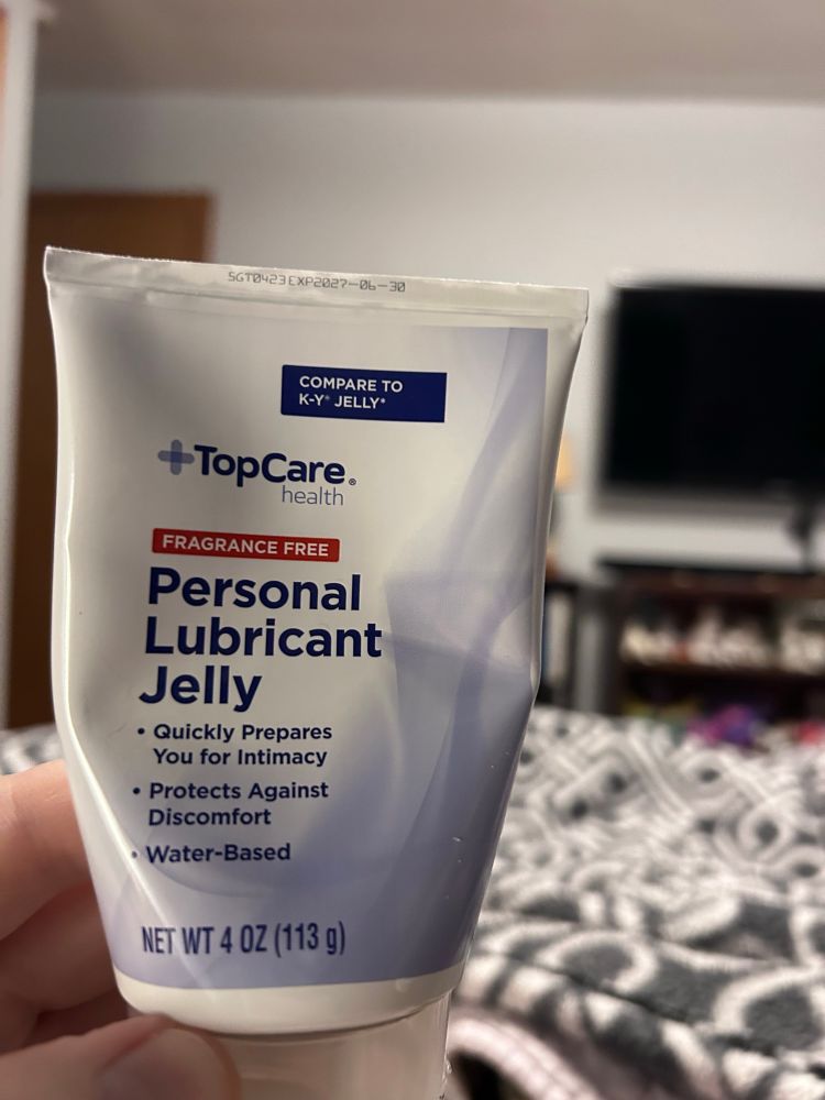 Lying in bed and almost non-visible woman holds a container of personal lubricant jelly close to the camera. You can only see the fingers and thumb of her left hand. This is top Care brand personal lubricant jelly. Beyond this image, you see a blanket on the bed and a TV on the wall in the background that is off.