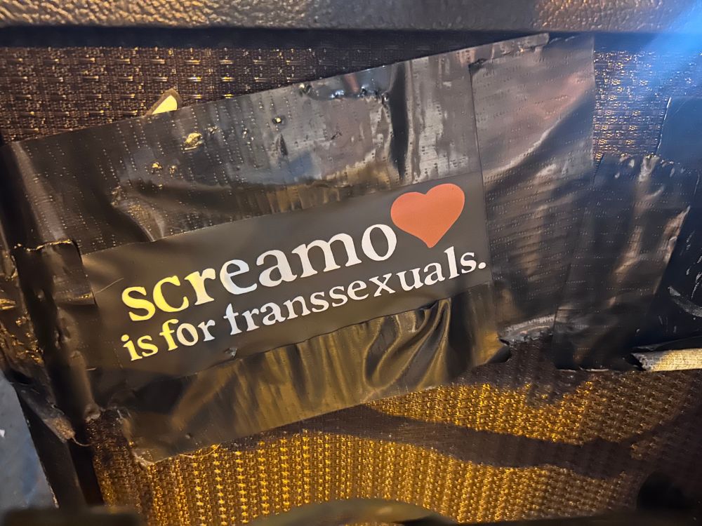 A close-up of a guitar ramp shows a sticker reading screamo is for transsexuals with a ❤️.