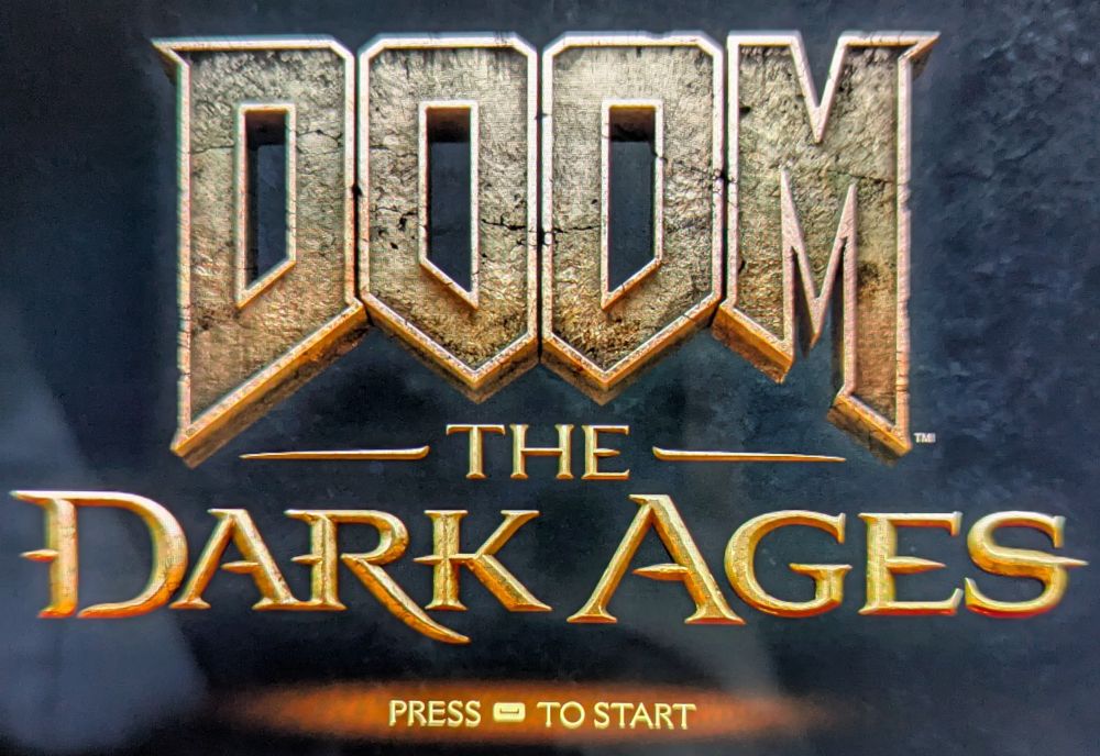 The logo from Doom: The Dark Ages as displayed on its main menu, with the iconic Doom logo in grey and gold tones as if made of stone and gold embossed letters with the subtitle beneath. It all sits on a background of dark grey stone. At the very bottom are the words 