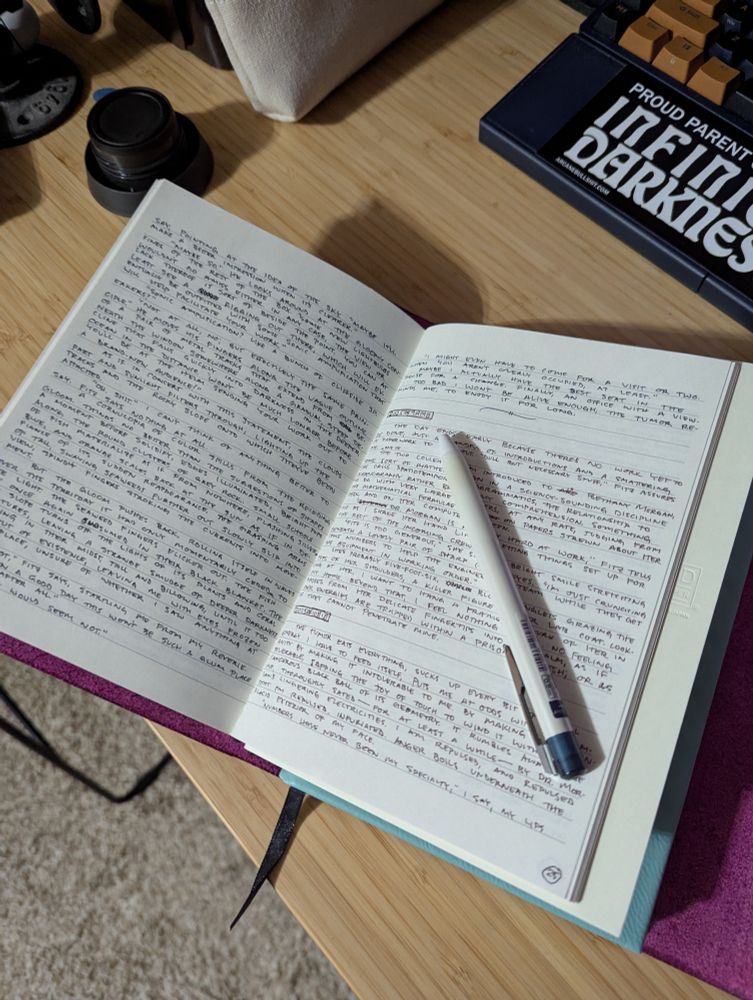 Michael J. Riser's journal (three journals in a maroon leather cover), open to a page with a lot of tiny serial-killer writing, an Ilmily pilot ballpoint in Nuance Back Blue sitting on top. A bamboo desk sits beneath. A keyboard above can be partially seen, with a bumper sticker on it that reads "proud parent of infinite darkness".
