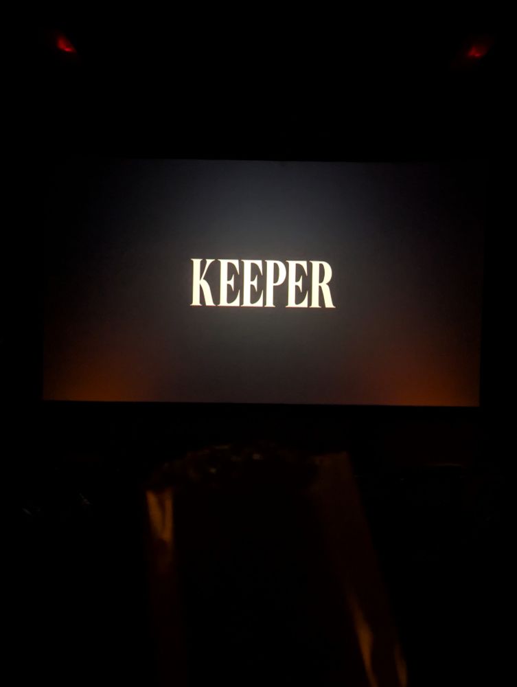 The title card for KEEPER, a horror film by Osgood Perkins, in a darkened theater with a bag of popcorn in front of it.
