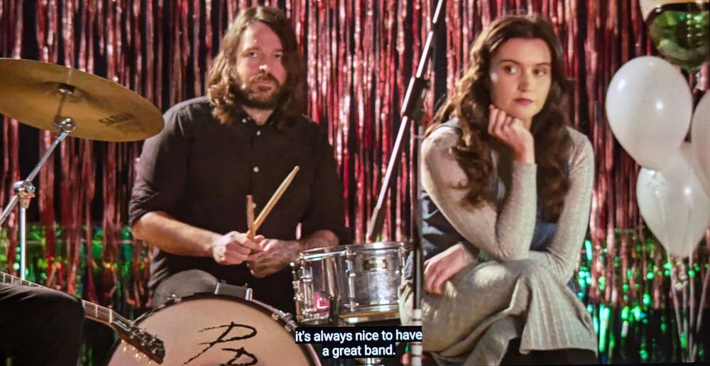 A still image from All You Need Is Death, showing one of the main characters sitting as part of a band at a wedding. She looks pensive, sitting in front of and next to a drummer, holding her head up with one hand, legs crossed. Shiny red streamers make a curtain behind them on the stage and there are white balloons to the right. The subtitles read, 