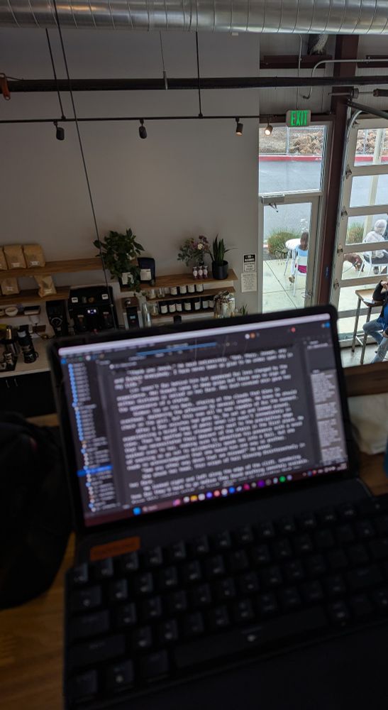 An image of horror author Michael J. Riser's laptop in a coffee shop. The words on the screen are out of focus, showing a busy field of Scrivener screens, while a cozy wall of coffee paraphernalia sits downstairs below, a view of the outside offered on some tall windows to the right.