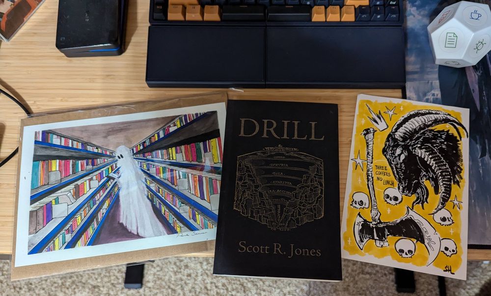 A photograph of horror author Michael J. Riser's writing desk on which two pieces of art and a book are displayed. On the left is a print of Paula Sanmarco's 