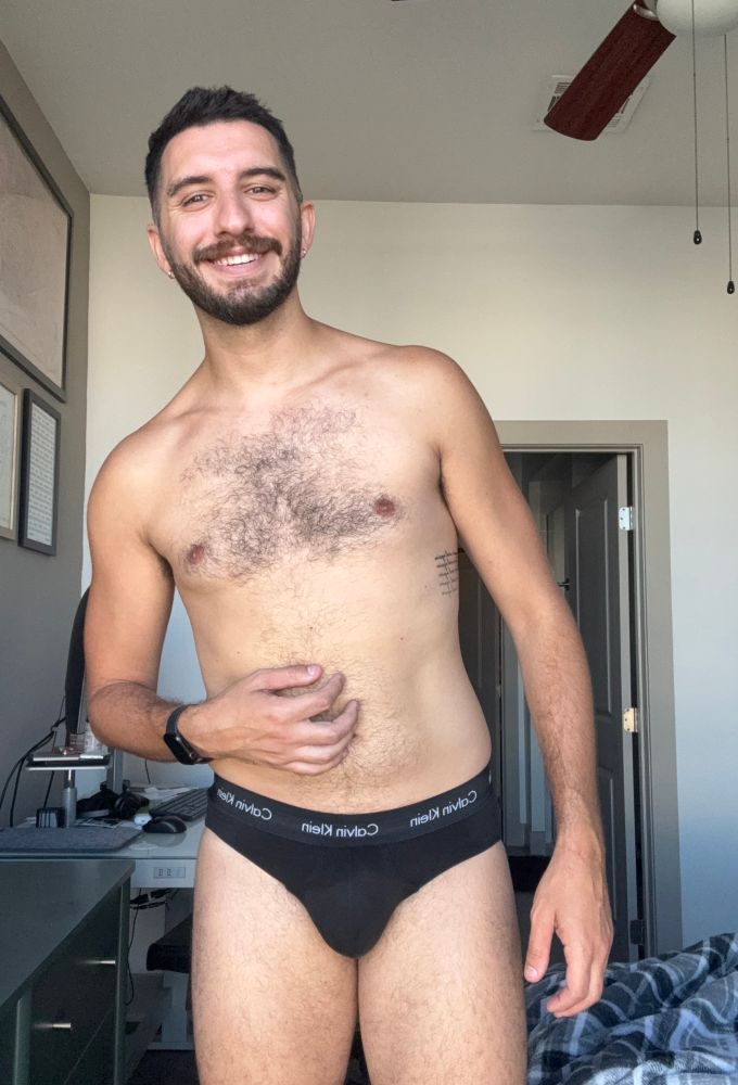 A picture of me standing in my bedroom in my black Calvin Klein briefs. Bed, sit/stand desk, framed art, and green dresser in the background. 