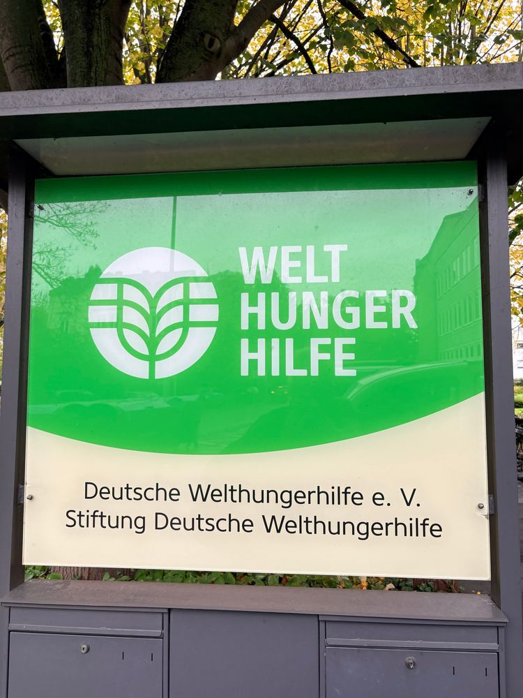 A picture of the logo of Welthungerhilfe in green and white. The picture is taken outside their office and a reflection of buildings can be seen in the glass sign that carries the logo. 