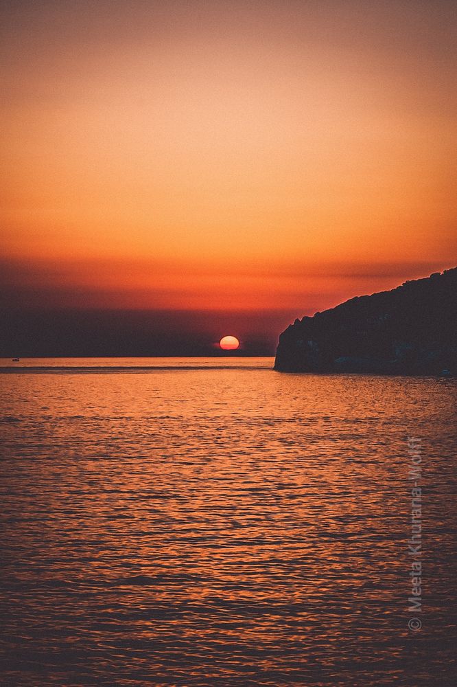 A vivid sunset over the sea in Pula, Croatia. The sun hovers just above the horizon, casting deep orange and red hues across the rippling water. A dark silhouette of a coastal cliff frames the right side, while the sky fades from burnt amber to dusky lavender, evoking a sense of quiet stillness. Image copyright Meeta K Wolff 