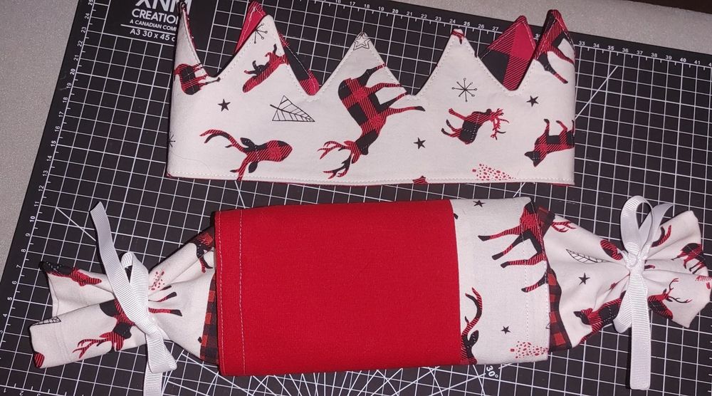 An image of a fabric reusable Christmas cracker crown with red plaid animals on a white background and a fabric Christmas cracker with patchwork fabric of the same red plaid animals on a white background, solid red and red plaid and ribbons to tie the ends. They are sitting on a black cm ruler cutting mat