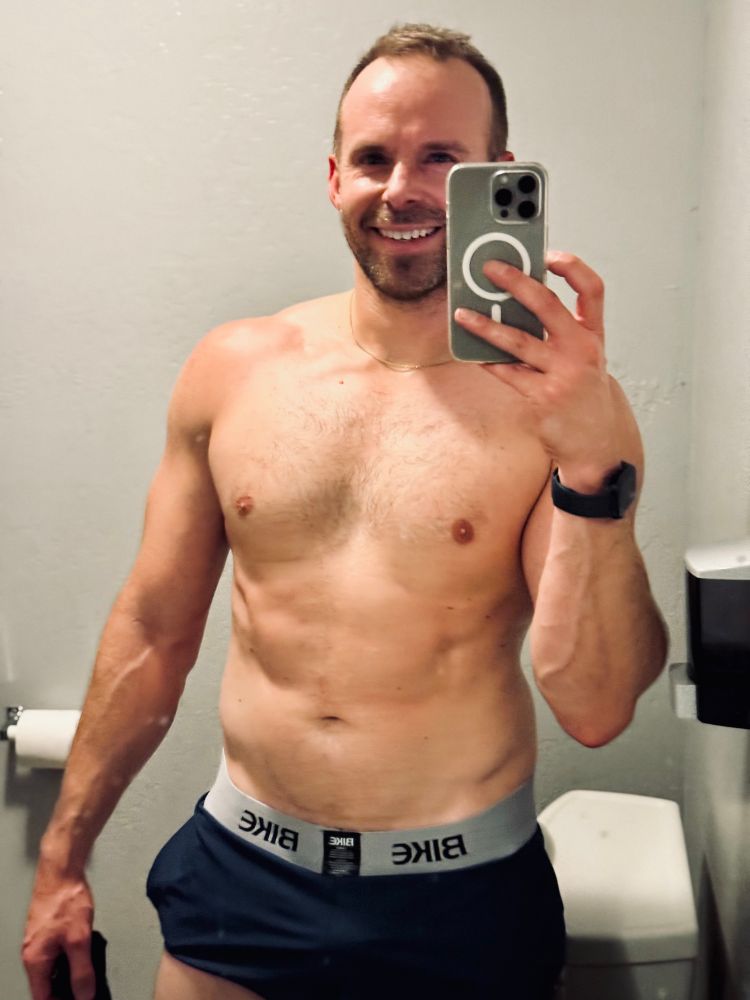 Shirtless man taking a mirror selfie in a small bathroom after a CrossFit workout, wearing BIKE hoochie daddy shorts