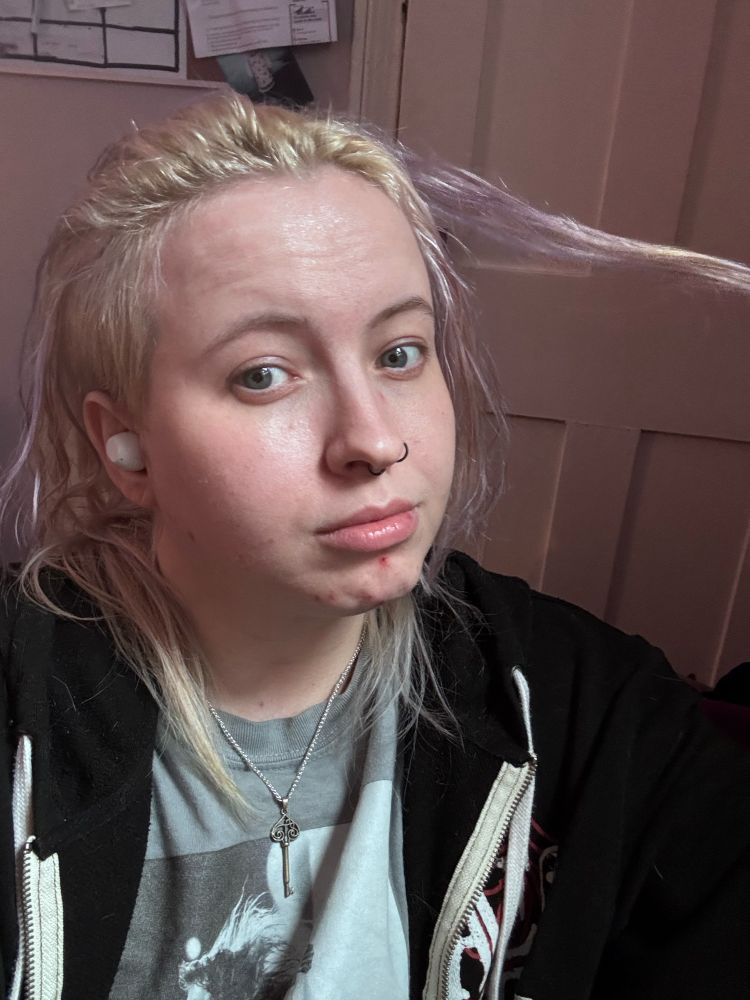 a white skinned person with white blond hair with blue eyes and a key necklace with a grey tee shirt and black and white hoodie on.