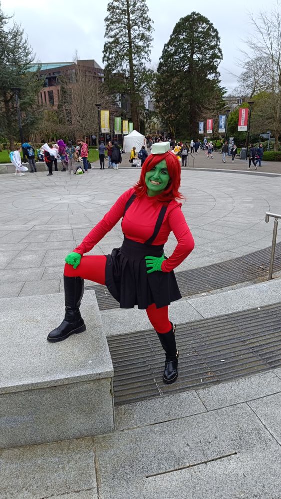 Photo of me in my Annie cosplay, with red long sleeve top, red tights, black dungaree-skirt, black boots, green gloves, red wig, green origami hat and green face paint.