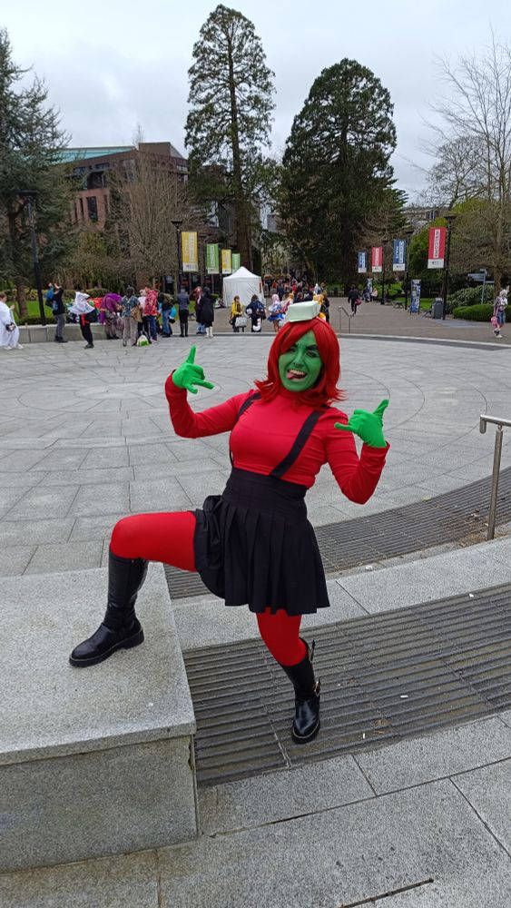 Photo of me in my Annie cosplay, with red long sleeve top, red tights, black dungaree-skirt, black boots, green gloves, red wig, green origami hat and green face paint.