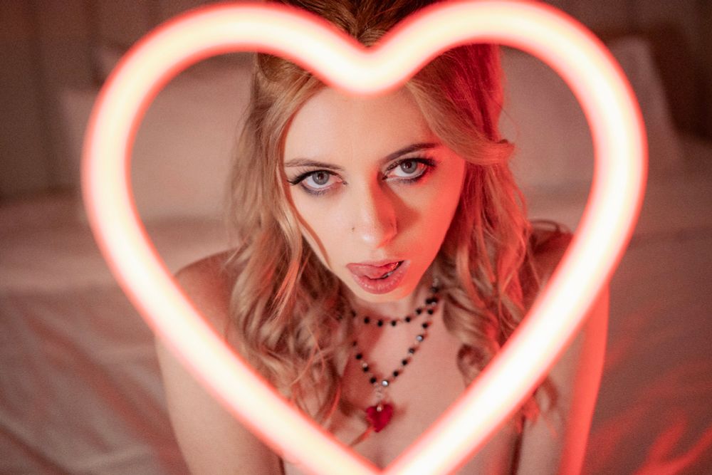 Portrait of eluna with an led heart close to the lens, framing her face. She is gently licking her lip and gazing into the camera. The vibe is very glamorous femme fatale with red lighting kissing her blonde hair, and she is ready to eat you. 