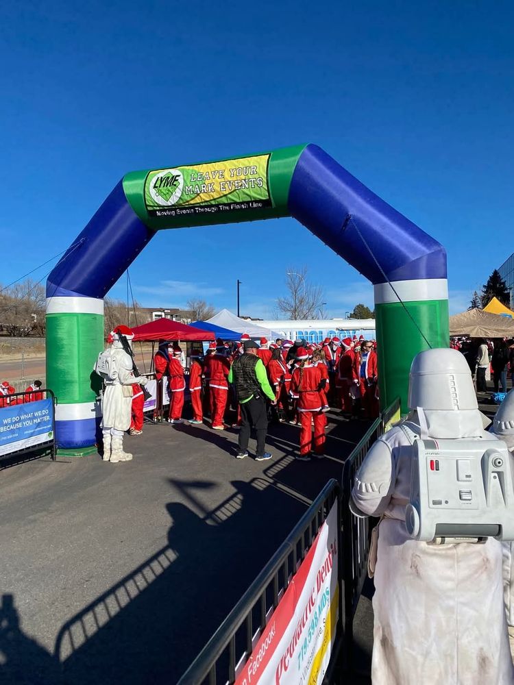 A photo of a large group of people all dressed as Santa's gathering for a 5K marathon in front of a green and blue blow-up arch. There are two, white Snowtroopers standing on either side of the path looking at the group.
